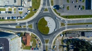 Drone view of roundabout at intersection of asphalt roads near modern buildings and parked cars in city district on sunny day